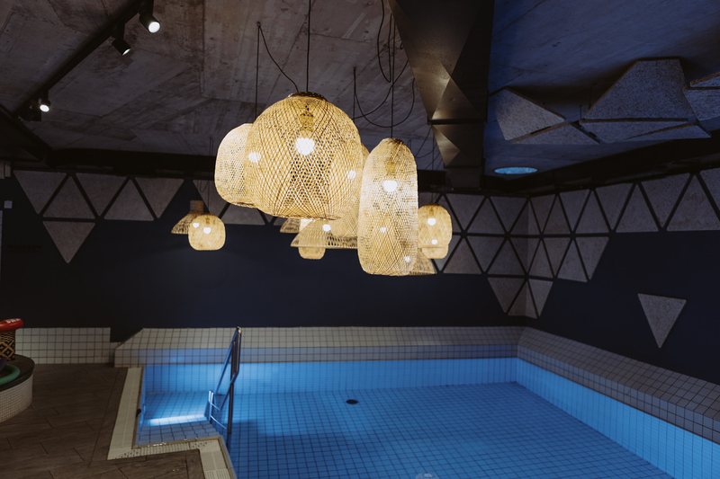 Pool im Alm-Spa mit Rattan-Hängeleuchten vor dunkelblauer Wand mit Dreiecksmuster.